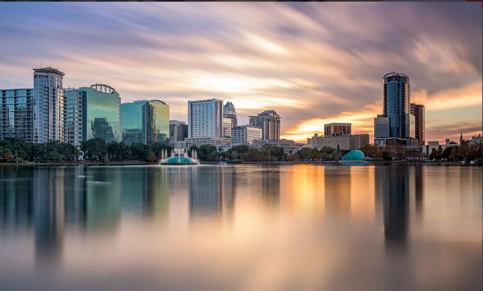 Aerial view of Lake Eola at sunset with Orlando skyline, purple and golden sky, sailboats on water, and surrounding parks and trees